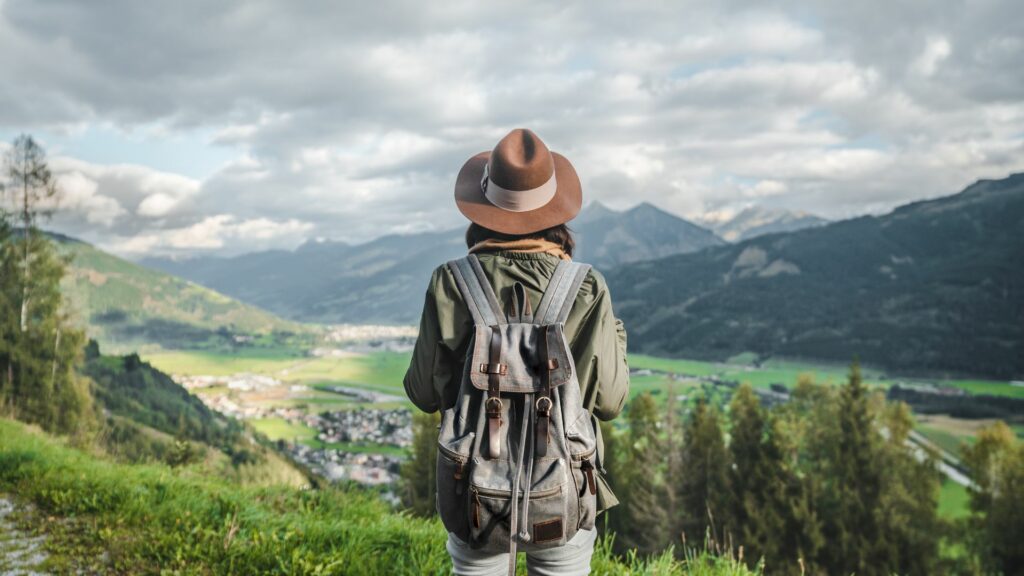 Imagem de uma jovem viajando pelas montanhas da Áustria. Ela está parada em um ponto de uma montanha, admirnado os campos verdes.
