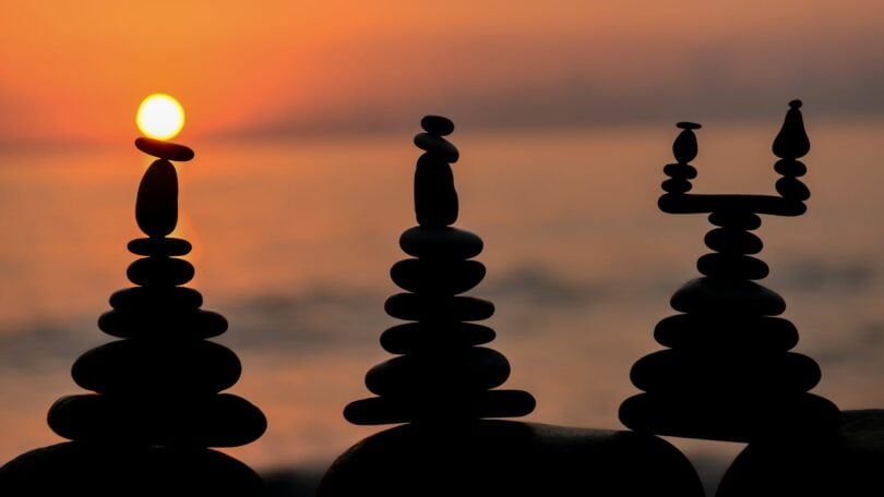 Imagem de 3 figuras feitas em pedra e ao fundo um lindo pôr do sol. A imagem traz o conceito de vida equilibrada e filosofia de vida.
