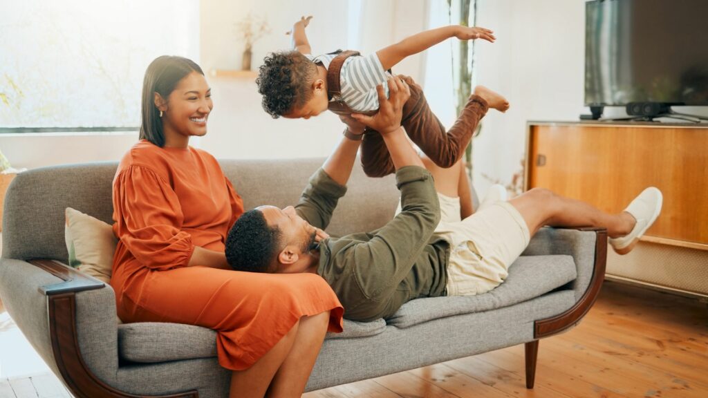 Imagem de uma família composta por pai, mãe e filho sentados em um sofá na sala de estar. Eles estão felizes e brincando o filho.
