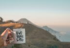 Imagem de fundo de uma montanha e em destaque a mão de uma mulher segurando uma caneca de porcelana onde esta escrityo: Viva sua vida, em inglês: Live Your Life.