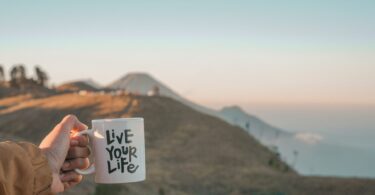 Imagem de fundo de uma montanha e em destaque a mão de uma mulher segurando uma caneca de porcelana onde esta escrityo: Viva sua vida, em inglês: Live Your Life.