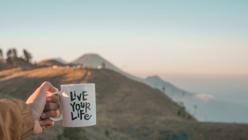 Imagem de fundo de uma montanha e em destaque a mão de uma mulher segurando uma caneca de porcelana onde esta escrityo: Viva sua vida, em inglês: Live Your Life.