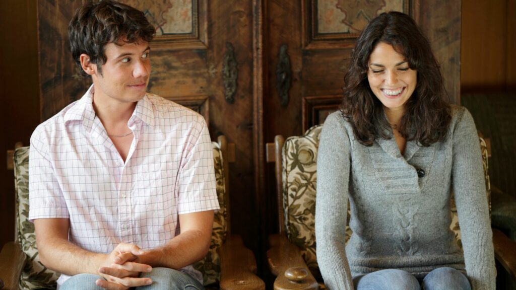 Imagem de um casal sentados e tentando estabelecer uma conversa. O homem olha para a mulher e ela está tímida, porém com um leve sorriso no rosto.
