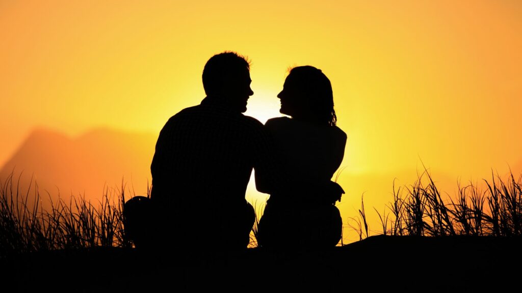 Imagem de um lindo pôr do sol e em destaque um casal sentando, conversando e de braços dados.
