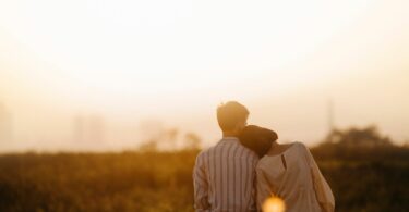 Imagem de um casal, homem e mulher, sentados olhando para o pôr do sol. Ela está com a cabeça encostada no ombro dele.