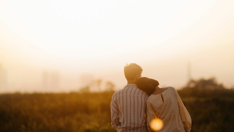 Imagem de um casal, homem e mulher, sentados olhando para o pôr do sol. Ela está com a cabeça encostada no ombro dele.