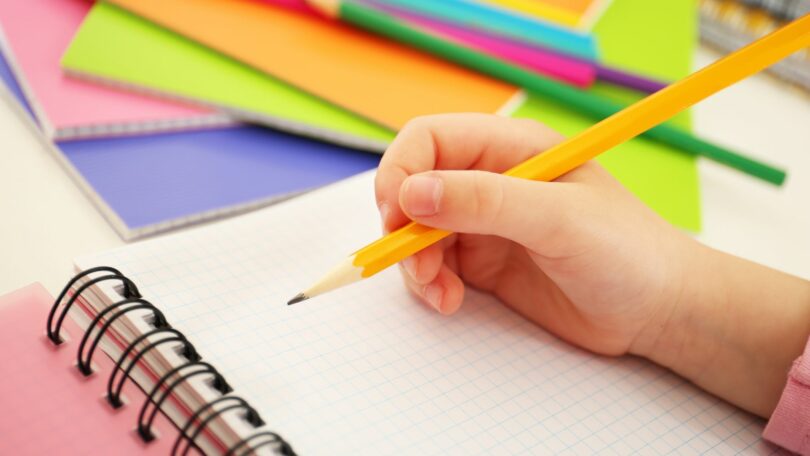 Imagem da mão de criança escrevendo em um caderno com um lápis.