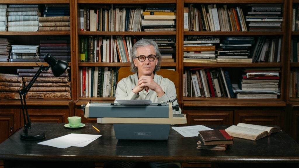 Imagem de um homem em uma sala cheio de livros. Ele está sentado em uma mesa, em frente à uma máquina de escrever, trazendo o conceito de uma vida intelectual.
