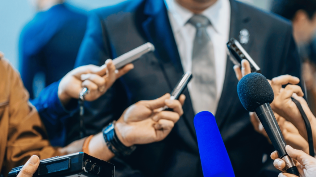 Na imagem, vários jornalistas estão ao redor de um político com seus microfones apontados para ele.