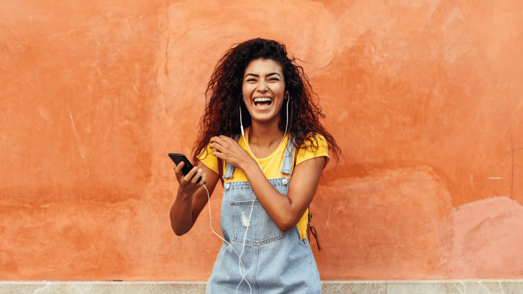 Na imagem, uma mulher negra e jovem está sorrindo e mexendo no celular com seus fones de ouvido.