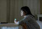 Na imagem, uma mulher asiática e jovem está no seu quarto assistindo televisão e segurando uma caneca.