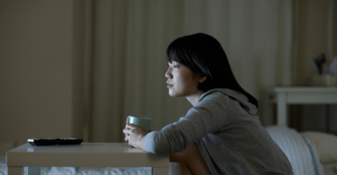 Na imagem, uma mulher asiática e jovem está no seu quarto assistindo televisão e segurando uma caneca.