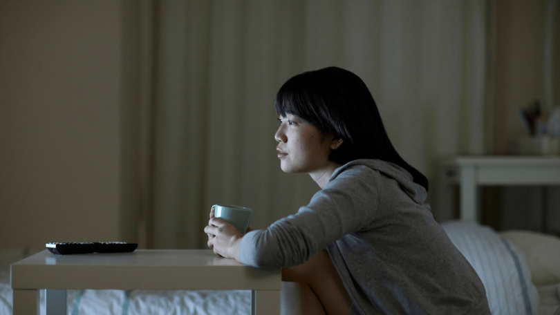 Na imagem, uma mulher asiática e jovem está no seu quarto assistindo televisão e segurando uma caneca.