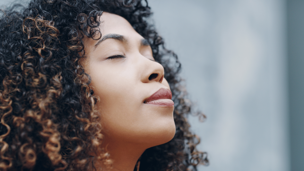 Na imagem, uma mulher negra e jovem está com a cabeça levantada, os olhos fechados e ela respira fundo.