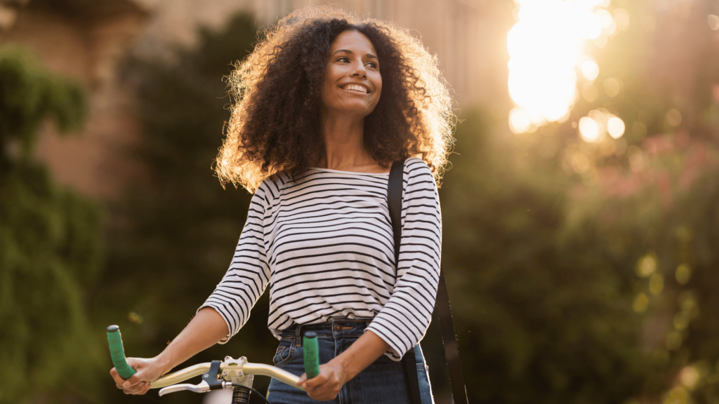 Na imagem, uma mulher negra e jovem está caminhando e segurando sua bicicleta. Ela olha para o lado e sorri.