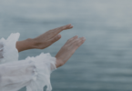 Na imagem, as mãos de uma mulher estão em destaque. Ela estende suas mãos no ar. Ao fundo, há o mar.