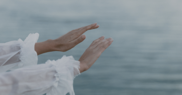 Na imagem, as mãos de uma mulher estão em destaque. Ela estende suas mãos no ar. Ao fundo, há o mar.