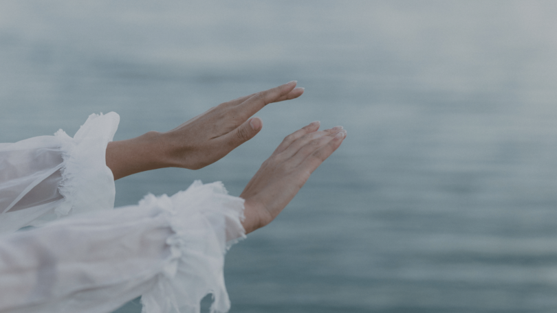 Na imagem, as mãos de uma mulher estão em destaque. Ela estende suas mãos no ar. Ao fundo, há o mar.