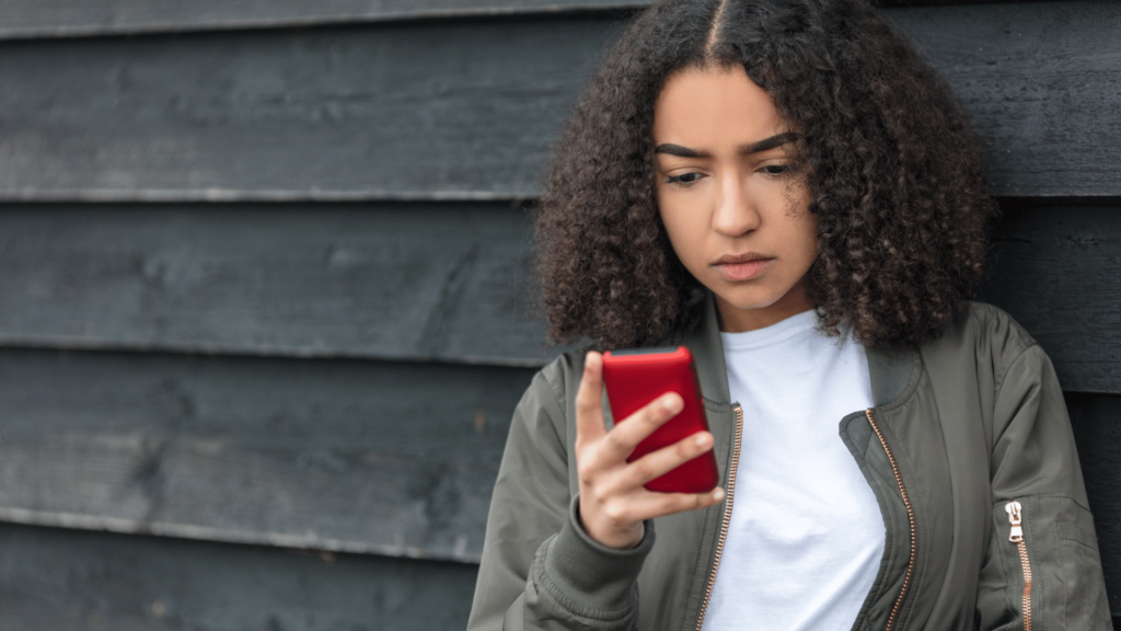 Uma mulher jovem e negra está checando e mexendo no celular. Ela está encostada em um portão em um ambiente aberto.