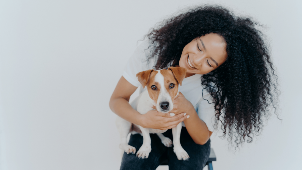 Uma mulher jovem e negra está sentada em um banco com seu cachorro no colo. Ela faz carinho no animal e sorri.