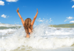 Uma mulher jovem e loira está entrando no mar e ela levanta os dois braços ao chocar-se com a onda.