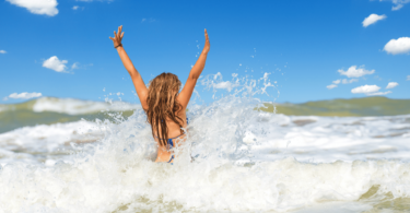 Uma mulher jovem e loira está entrando no mar e ela levanta os dois braços ao chocar-se com a onda.