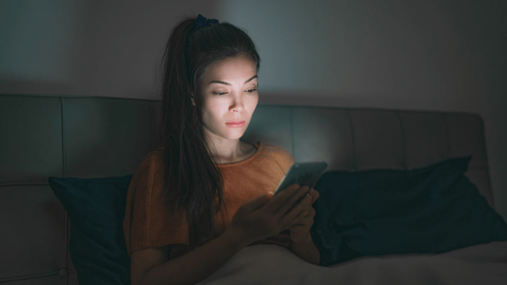 Uma mulher jovem e asiática está sentada na sua cama e mexendo no celular durante a noite.