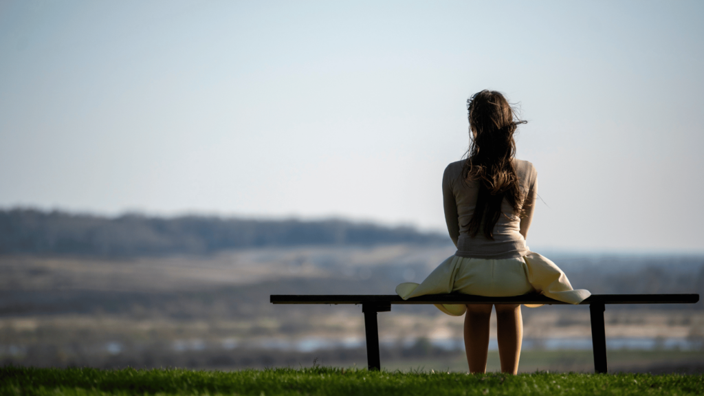 Uma mulher está de costas para a imagem e sentada em um banco ao ar livre. Ela está contemplando a paisagem.