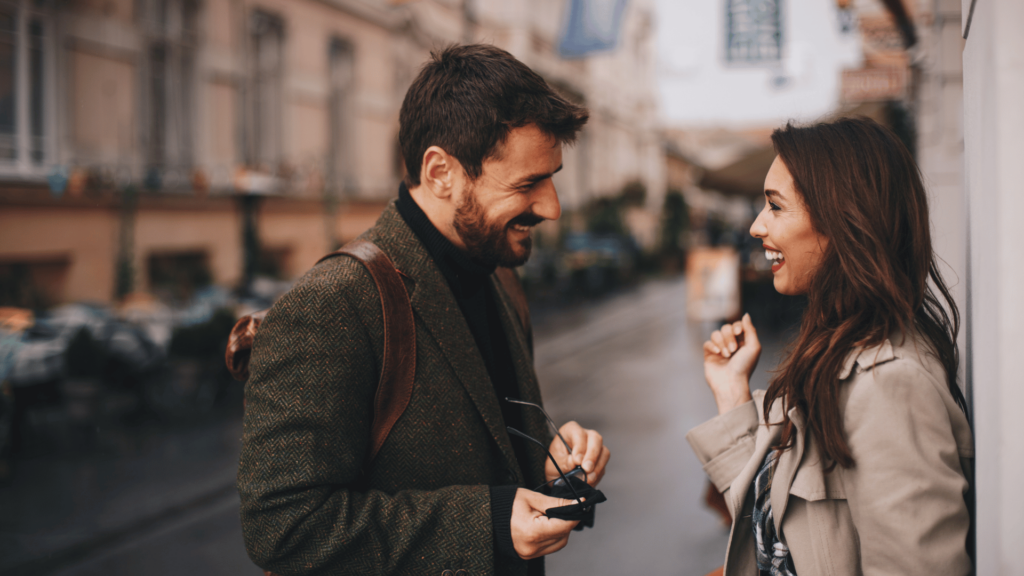 Um homem e uma mulher estão sorrindo um para o outro e flertando. Eles estão em uma rua.