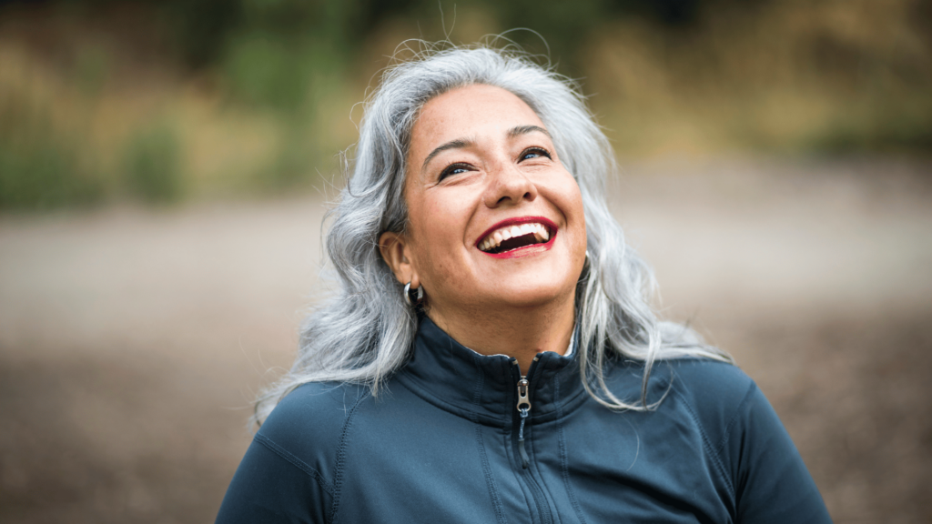 Imagem de uma mulher, por volta dos 40 ou 50 anos, sorrindo e olhando para cima. Ela está ao ar livre e um ambiente de natureza.