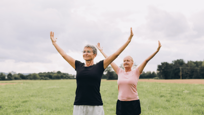 Imagem de duas mulheres, por volta dos 50 ou 60 anos, praticando Tai Chi ao ar livre em um parque.