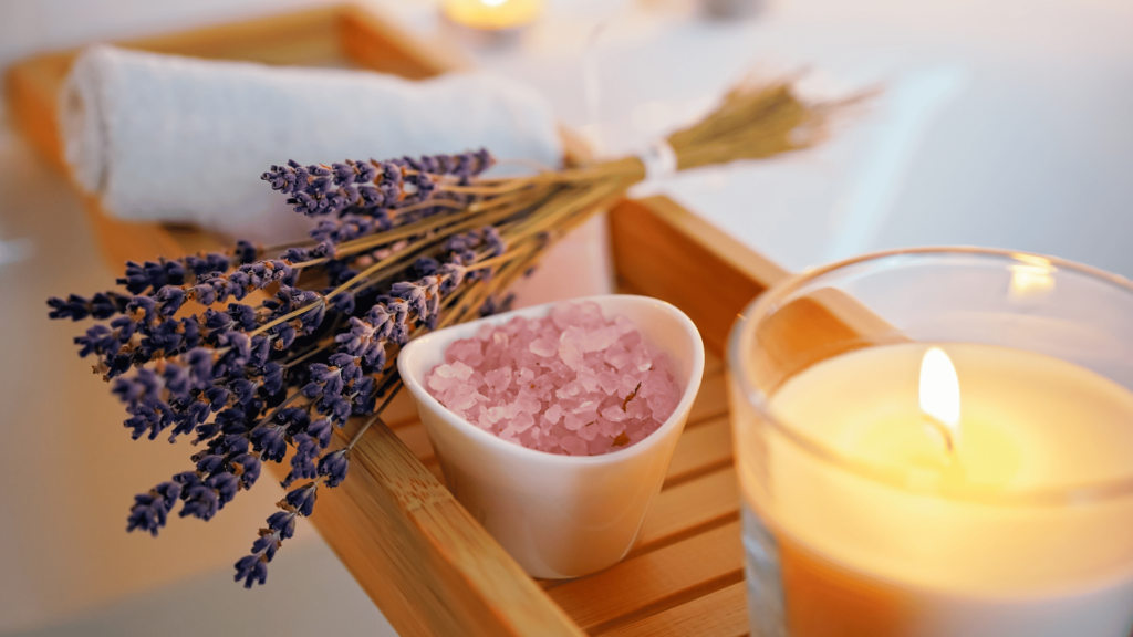 Um banho está sendo preparado com alguns objetos, como: lavanda, sal grosso, vela e uma toalha. Ao fundo, há uma banheira.