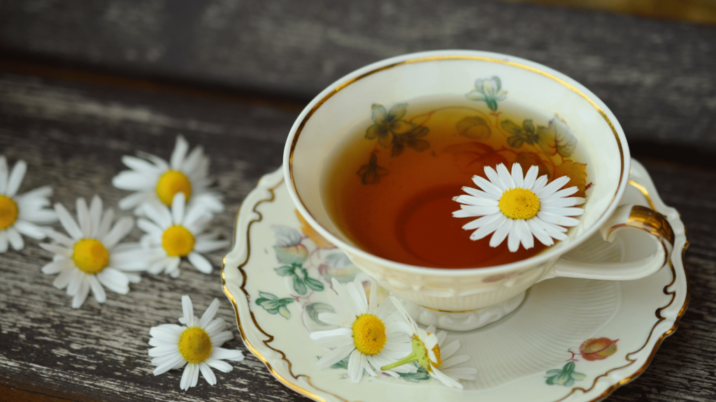 Uma xícara com chá de camomila está no foco da imagem. Há uma flor de camomila dentro do recipiente e mais flores fora.
