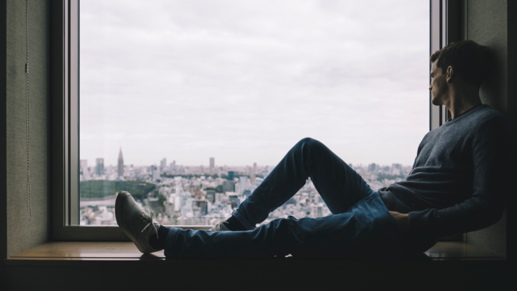 Um homem está sentado no parapeito de uma janela, olha pela janela e visualiza a cidade.