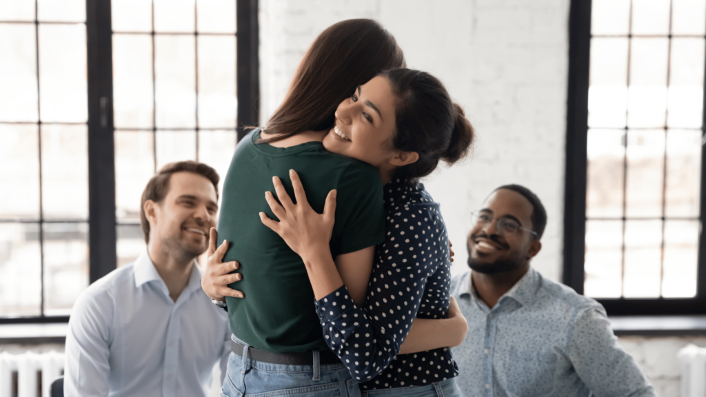 Uma mulher abraça outra mulher e sorri. Atrás delas, estão sentados dois homens que também estão sorrindo. Eles estão em um ambiente de trabalho.