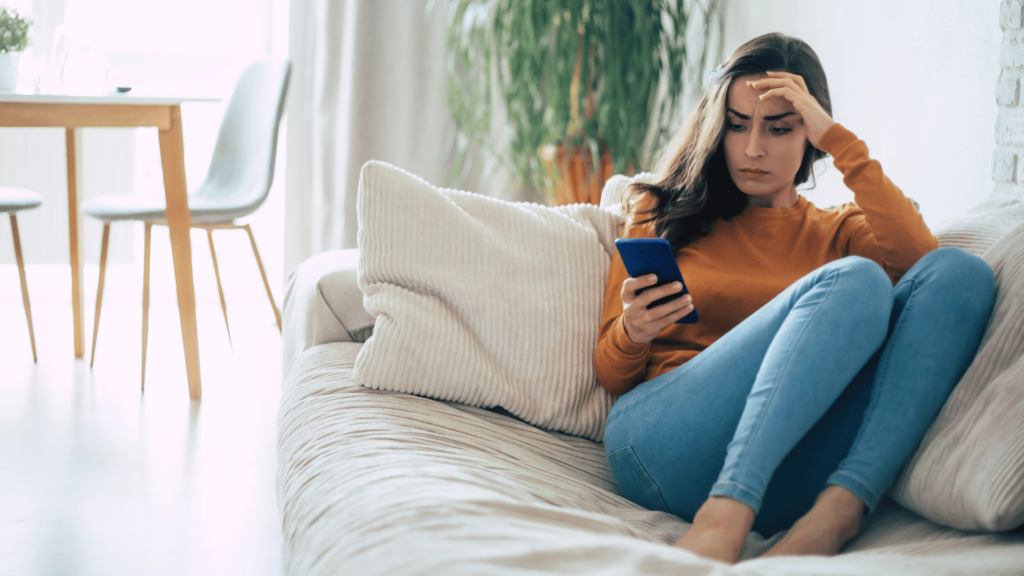 Uma mulher está sentada no seu sofá e está mexendo no celular. Ela tem a expressão de frustração no rosto.