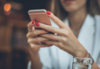 As mãos de uma mulher segurando e mexendo no seu celular estão no foco da imagem.