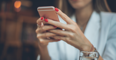 As mãos de uma mulher segurando e mexendo no seu celular estão no foco da imagem.