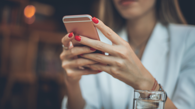 As mãos de uma mulher segurando e mexendo no seu celular estão no foco da imagem.