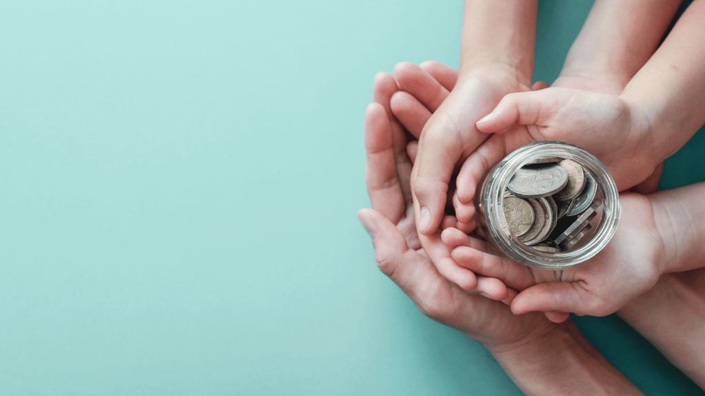 As mãos de uma mãe, um pai e um filho (criança) estão umas sobre as outras segurando um pote com dinheiro dentro, indicando que são as economias da família.