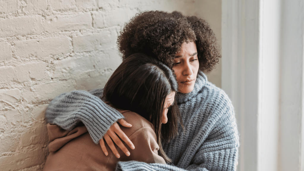 Duas mulheres estão se abraçando. Uma está dando apoio para a outra.