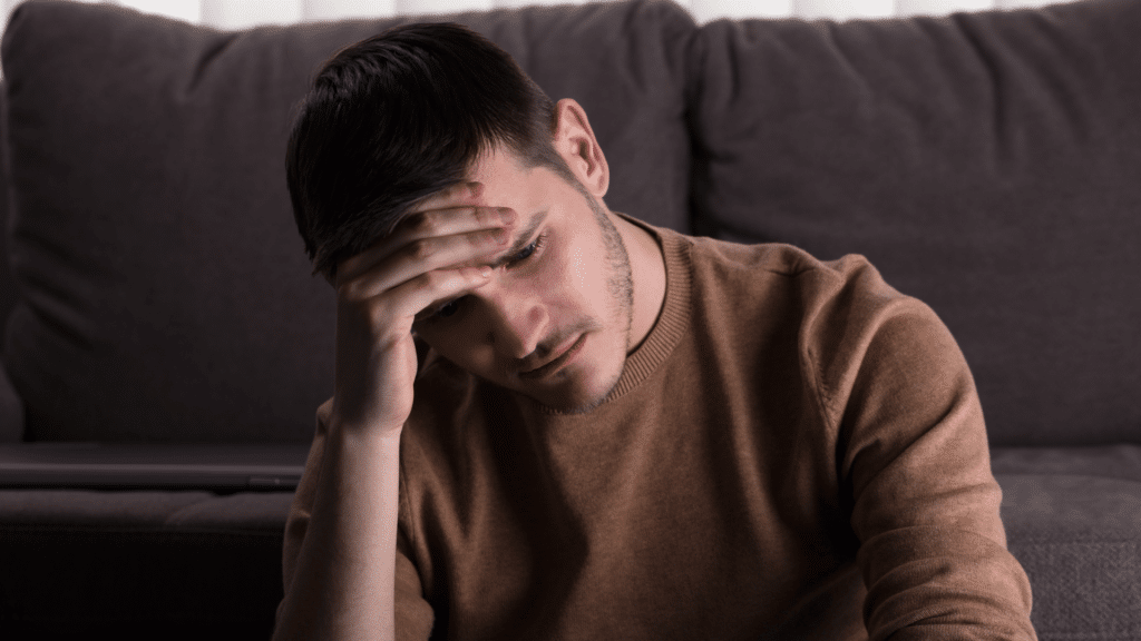 Um homem triste e cabisbaixo está sentado na frente de um sofá com uma mão na cabeça.