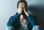 Um homem triste ou preocupado está sentado no chão e encostado em uma parede. Ele coloca as duas mãos no rosto.