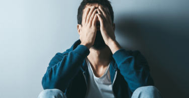 Um homem triste ou preocupado está sentado no chão e encostado em uma parede. Ele coloca as duas mãos no rosto.