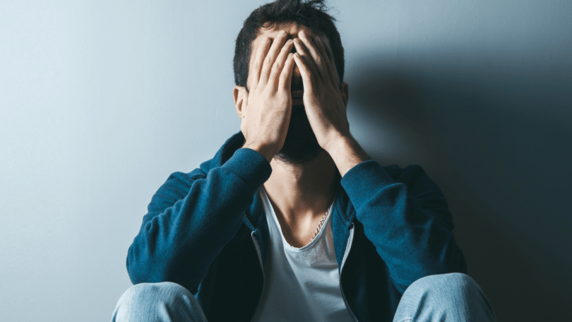Um homem triste ou preocupado está sentado no chão e encostado em uma parede. Ele coloca as duas mãos no rosto.