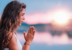 Uma mulher que junta as duas mãos e medita ao ar livre está no foco da imagem.