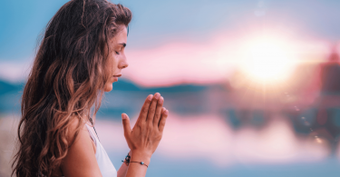 Uma mulher que junta as duas mãos e medita ao ar livre está no foco da imagem.
