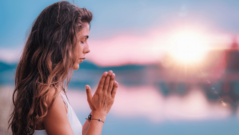 Uma mulher que junta as duas mãos e medita ao ar livre está no foco da imagem.