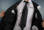 Um homem de negócios vestido com roupas formais está no foco da imagem. Ele abre seu terno e mostra várias notas de dólares espalhadas pelos seus bolsos.