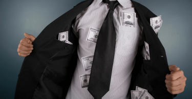 Um homem de negócios vestido com roupas formais está no foco da imagem. Ele abre seu terno e mostra várias notas de dólares espalhadas pelos seus bolsos.